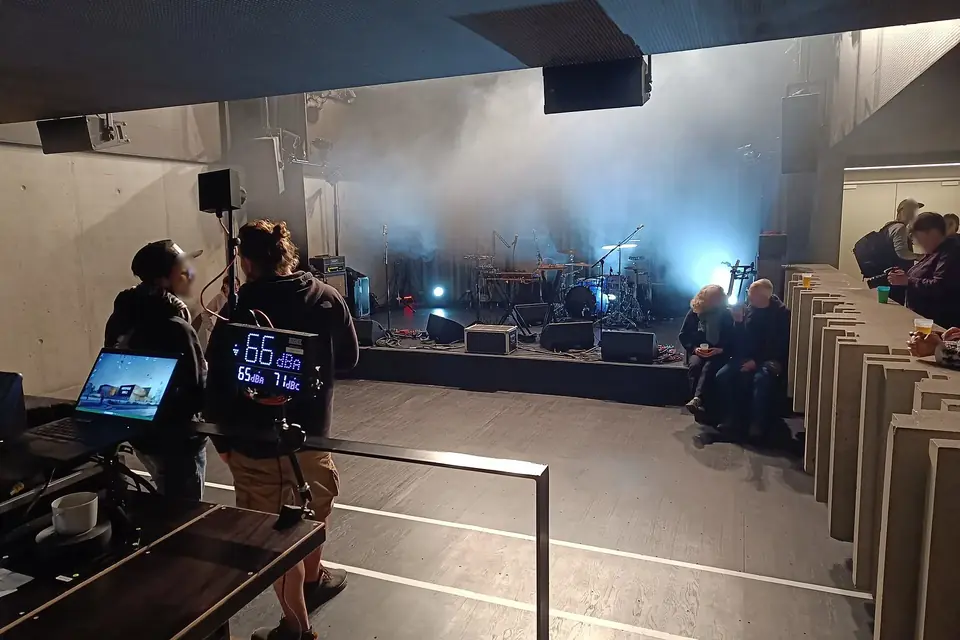 Une petite salle de concert sombre avec une scène éclairée en bleu, une régie au premier plan et quelques spectateurs