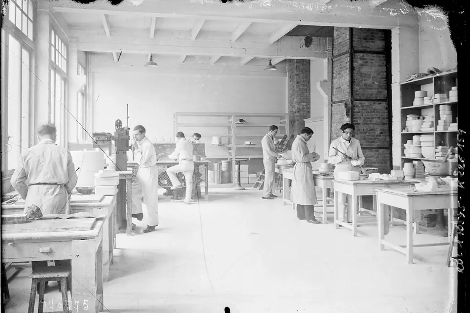 Photographie ancienne en noir et blanc d'un vaste atelier de céramique, avec des établis, des étagères de pièces et des élèves au travail