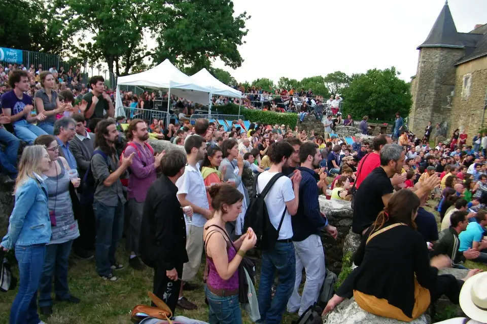 Une foule nombreuse rassemblée en plein air dans un parc arboré, devant des tentes blanches et une tour de manoir