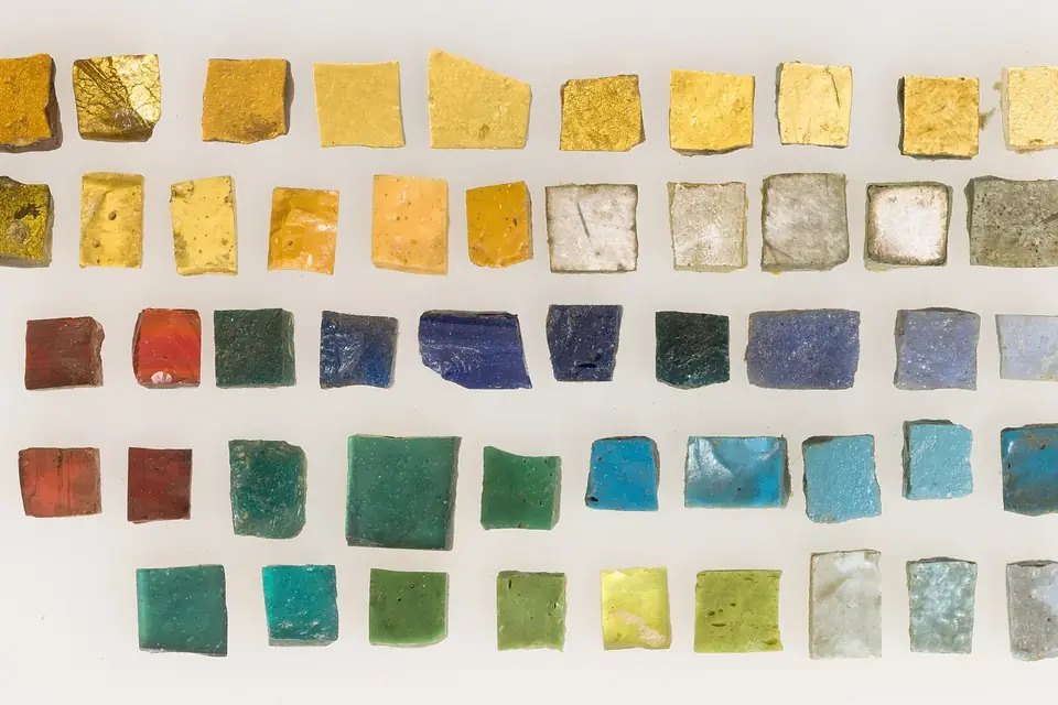 Des tesselles de mosaïque en verre, dorées, vertes, bleues et brunes, alignées sur un fond blanc